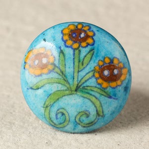 Puede incluir: Perilla de armario redonda de cerámica turquesa con diseño floral. La perilla presenta tres girasoles amarillos y marrones con tallos y hojas verdes. El fondo es azul claro moteado.