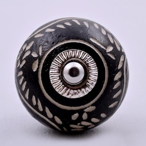May include: A round cabinet knob featuring a matte black finish with carved white leaf patterns. The centre of the knob has a circular silver-toned metal plate with a ridged edge and a smooth, polished silver-toned metal bolt head in the middle.