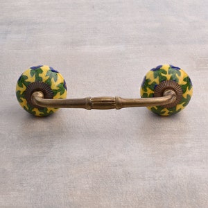 May include: A brass drawer pull with two round ceramic knobs. The knobs are yellow with green and blue floral designs.