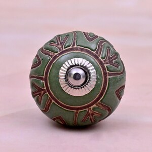 Op de afbeelding: Een ronde keramische meubelknop in een gedempte saliegroene kleur met een donkerbruin reliëfpatroon van botanische vormen. Het midden van de knop heeft een cirkelvormig zilverkleurig metalen onderdeel met een geribbelde rand en een gladde koepel.