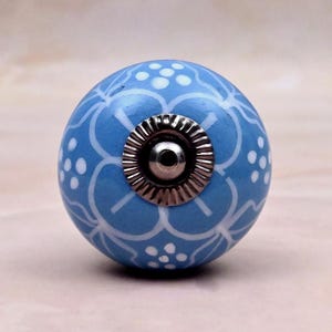 Op de afbeelding: Een ronde keramische meubelknop in lichtblauw met een wit handgeschilderd bloemmotief met kleine stippen. Het midden van de knop heeft een cirkelvormig zilverkleurig metalen beslag met een geribbelde rand en een centraal schroefgat.
