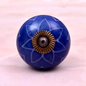 Op de afbeelding: Een bolvormige blauwe meubelknop met een wit geëtst bloemmotief over het oppervlak. Het midden van de knop is versierd met een cirkelvormige, geribbelde messingkleurige metalen plaat en een klein, donker centraal schroefbevestigingspunt.