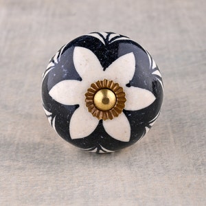 May include: A white ceramic knob with a black floral design and a gold center. The knob is round and has a smooth surface.