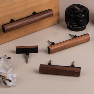 Könnte beinhalten: Schrankgriffe aus Holz mit dunkelbraunem Finish. Die Griffe sind rechteckig mit schwarzem Sockel und silbernen Schrauben. Ein Griff ist an einer Holzoberfläche befestigt. Ein Canon-Objektiv befindet sich im Hintergrund.