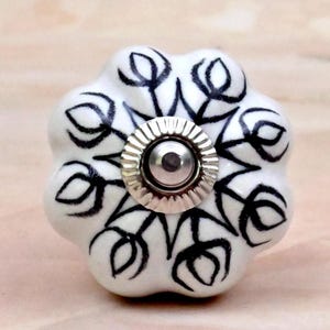 Op de afbeelding: Een witte keramische meubelknop met een geschulpte, bloemachtige vorm. Het oppervlak is voorzien van een met de hand geschilderd zwart lijnpatroon dat lijkt op gestileerde tulpen. Een cirkelvormig, zilverkleurig metalen onderdeel zit in het midden.