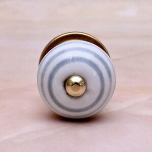 May include: A round ceramic cabinet knob featuring a white base with concentric light grey stripes. The centre of the knob is accented with a small, polished gold-tone metal stud, and the base of the knob is finished in a matching gold-tone metal.