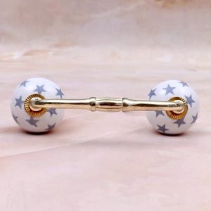 May include: A drawer pull featuring a gold-toned metal handle connected to two spherical white ceramic knobs. Each knob is decorated with a repeating pattern of small grey stars. The metal handle has a smooth, segmented design with ribbed accents.