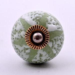 Peut inclure: Un bouton de meuble rond en céramique présentant un motif marbré vert pâle et blanc avec un design en forme d'étoile. Le centre du bouton est orné d'une pièce métallique dorée circulaire et striée ainsi que d'une petite vis argentée.