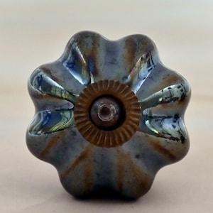 May include: A flower-shaped cabinet knob with a reflective, iridescent finish in shades of blue, grey, and brown. The centre features a circular, ridged brass-toned metal plate with a small, protruding screw head in the middle of the design.