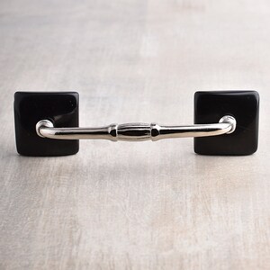 May include: A cabinet handle featuring a polished silver-toned metal bar with a central decorative detail, connected to two square, glossy black base plates. The hardware has a smooth, reflective finish and a horizontal orientation.