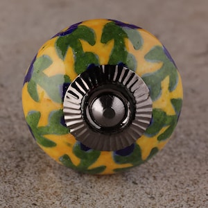 May include: A decorative cabinet knob with a yellow, green, and blue floral pattern. The knob has a dark metal base and a round, textured design. This unique hardware adds a pop of color and style to any furniture piece.