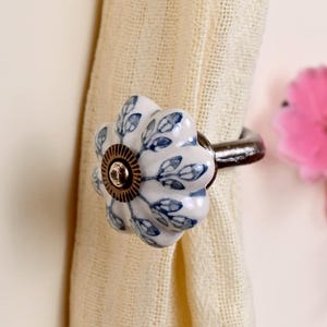 May include: A ceramic curtain tieback featuring a white flower-shaped knob with a blue leaf pattern. The center of the knob has a circular bronze-toned metal accent. A dark metal hook extends from the back of the knob to secure the fabric.