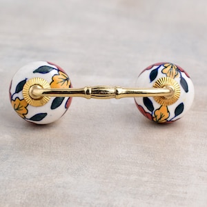 Könnte beinhalten: Ein weißer Keramik-Schubladengriff mit einer goldenen Metallstange, die zwei runde Knöpfe verbindet. Die Knöpfe sind mit einem floralen Muster in Rot-, Gelb- und Blautönen verziert.