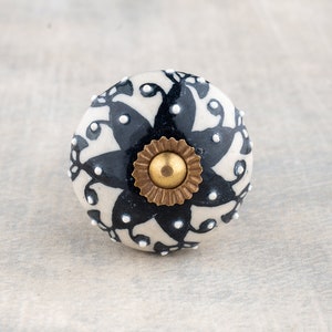 May include: White ceramic knob with a black floral design and gold accents. The knob has a raised edge and a small, round knob in the center.