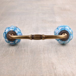 Puede incluir: Un tirador de cajón de cerámica azul y blanco con una barra de latón que conecta los dos pomos. Los pomos tienen forma de flor con un diseño en relieve.