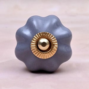 May include: A grey ceramic cabinet knob shaped like a flower with six rounded petals. The center of the knob features a circular gold-colored metal accent with a fluted edge and a smooth, polished gold-colored dome in the very middle.