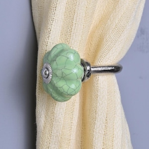 May include: A light green ceramic curtain tieback with a crackle finish, attached to a silver-toned metal ring. The tieback is holding a cream-colored curtain. The ceramic piece has a round shape with a small silver accent in the center.