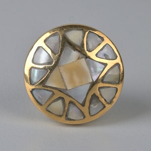 May include: A round, gold-toned knob with a geometric design. The knob is inlaid with mother-of-pearl pieces in a variety of shapes and sizes.