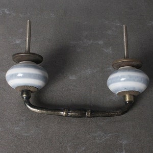 May include: A white and gray striped ceramic drawer pull with a brass finish. The pull has a curved shape and is attached to a metal rod.
