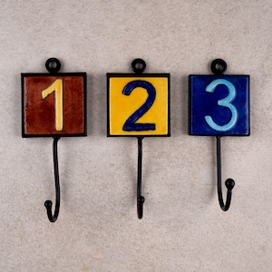 May include: Set of three decorative wall hooks. Each hook features a square tile with a number: 1 in yellow, 2 in blue, and 3 in teal. The tiles are set in black frames with a black hook below. The hooks are designed for hanging items.