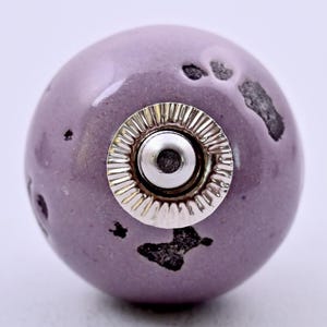 Puede incluir: Un pomo esférico de cerámica para muebles en color lavanda suave con un acabado brillante. La superficie presenta manchas irregulares de textura gris oscuro. Una base metálica circular de tono plateado con un borde estriado está centrada.