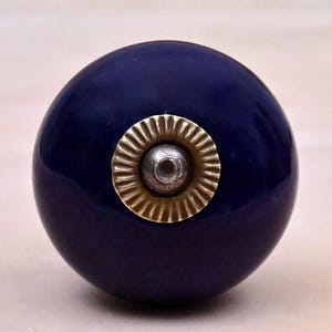 May include: A spherical dark blue cabinet knob with a smooth, glossy finish. The center features a circular, metallic gold-toned plate with a ribbed, sunburst-like texture surrounding a small, raised silver-colored metal bolt head.