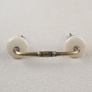 Pode incluir: Uma maçaneta de armário de cerâmica branca com uma pega de latão. A maçaneta é redonda e tem um acabamento liso. A pega é curva e tem um design simples.