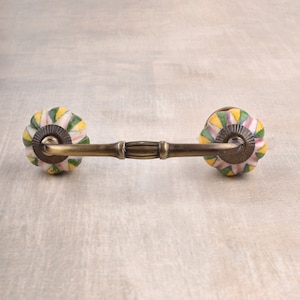 May include: A decorative drawer pull with a brass finish and two colourful ceramic knobs. The knobs feature a yellow, green and pink geometric pattern.