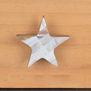 Może przedstawiać: Biały, gwiazdkowaty gałka do szafki wykonana z perłowej muszli. Gałka jest zamontowana na jasnobrązowej powierzchni drewnianej.