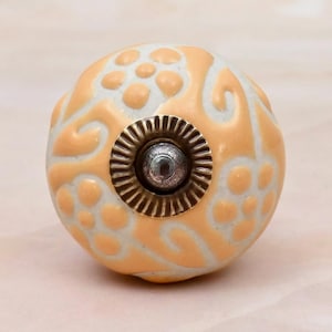 May include: A round ceramic cabinet knob featuring a light peach-coloured floral pattern embossed on a white background. The centre of the knob has a metallic, fluted circular plate with a small, dark, rounded metal stud protruding from the middle.