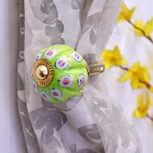 May include: A decorative curtain tieback with a bright green, round ceramic bead. The bead features a pattern of pink and white dots and a gold-colored metal accent. The tieback is holding a sheer, patterned curtain.