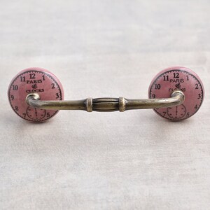 以下が含まれることがあります： 金色の金属製の腕が付いたピンクのセラミック製の引き出しの取っ手。セラミック製のノブには、「Paris Clocks」という文字が書かれた時計の文字盤があります。