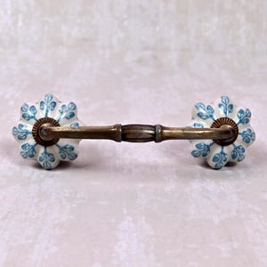 May include: A drawer pull featuring a horizontal antique brass metal handle connected to two white ceramic knobs. Each knob has a scalloped, flower-like shape and is decorated with a repeating blue botanical leaf pattern across the surface.
