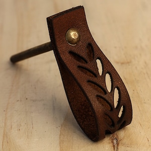 May include: Brown leather cabinet pull with a leaf design and a brass screw. The pull has a decorative brass rivet.