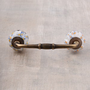 May include: A decorative cabinet pull with a brass finish and two white ceramic knobs. The knobs are adorned with a pattern of yellow and blue floral designs. The pull is designed for furniture and home decor.