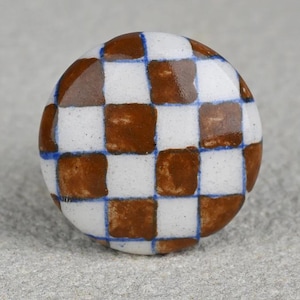 May include: A round, ceramic cabinet knob with a brown and white checkerboard pattern. The squares are outlined with thin blue lines. The knob is a decorative element for furniture and home decor.