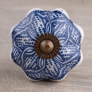 May include: A flower-shaped ceramic cabinet knob featuring a white base with a detailed blue lattice and floral pattern. The centre of the knob is accented with a circular, scalloped-edge bronze-toned metal hardware piece with a central dome.