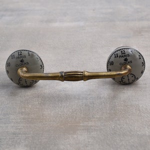 以下が含まれることがあります： 装飾的なデザインが施されたアンティーク調の真鍮製のキャビネットプル2個セット。各プルには、丸い磁器製のノブが付いており、時計の文字盤のデザインで「Paris Clocks」と表示されています。