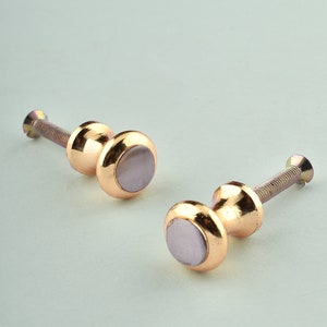 May include: Two gold-toned cabinet knobs with a round, slightly raised, off-white center. The knobs have a screw on the back for attaching to a cabinet door.
