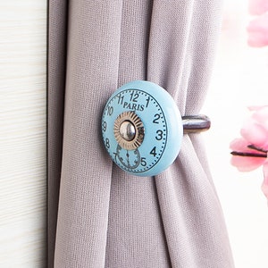 May include: A light blue ceramic curtain tieback with a clock face design. The clock face has black Roman numerals and the word "PARIS" printed on it. The tieback is attached to a metal hook.