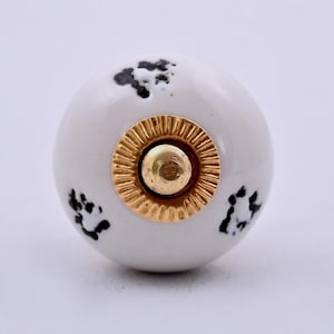 Op de afbeelding: Een ronde witte keramische meubelknop met een verspreid zwart spikkelpatroon. Het midden van de knop is versierd met een cirkelvormig goudkleurig metalen beslag met een geribbelde, getextureerde rand rondom een gladde centrale pin.
