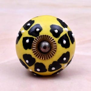 Op de afbeelding: Een ronde keramische meubelknop met een heldergele basiskleur en een zwart luipaardprintpatroon. Het midden van de knop heeft een goudkleurige metalen cirkelvormige plaat met een kleine zilverkleurige schroefkop in het midden.