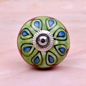 Op de afbeelding: Een ronde keramische meubelknop met een lichtgroene basis en een herhalend patroon van donker omrande druppelvormen met blauwe centra. Het midden van de knop is voorzien van een zilverkleurig metalen onderdeel met een geribbelde rand.