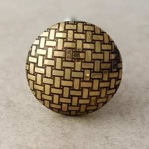 Puede incluir: Un pomo de armario redondo de color dorado con un patrón tejido.