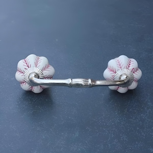 May include: White ceramic drawer pull with a pink floral design and a silver metal bar connecting the two sides.