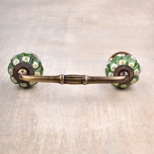 Pode incluir: Uma puxador de gaveta de cerâmica verde e branca com uma barra de latão que conecta as duas maçanetas. As maçanetas têm um padrão floral.