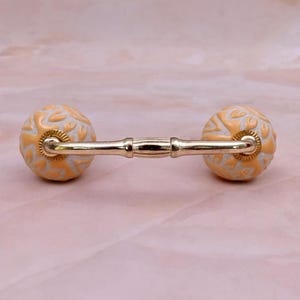May include: A drawer pull featuring two spherical knobs with a light orange and white embossed floral pattern, connected by a polished gold-tone metal bar. The hardware has a central decorative band and fluted circular bases at each connection point.