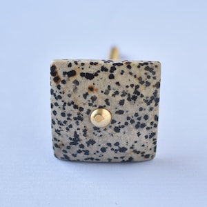 Op de afbeelding: Een vierkante knop met een gespikkeld patroon in zwart, wit en bruin. De knop heeft een gouden metalen accent in het midden.