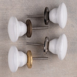 May include: Four white ceramic cabinet knobs with smooth, rounded tops and tapered bases. Each knob features a threaded metal bolt and a circular metal backplate in varying finishes, including dark bronze, polished silver, and brushed brass tones.