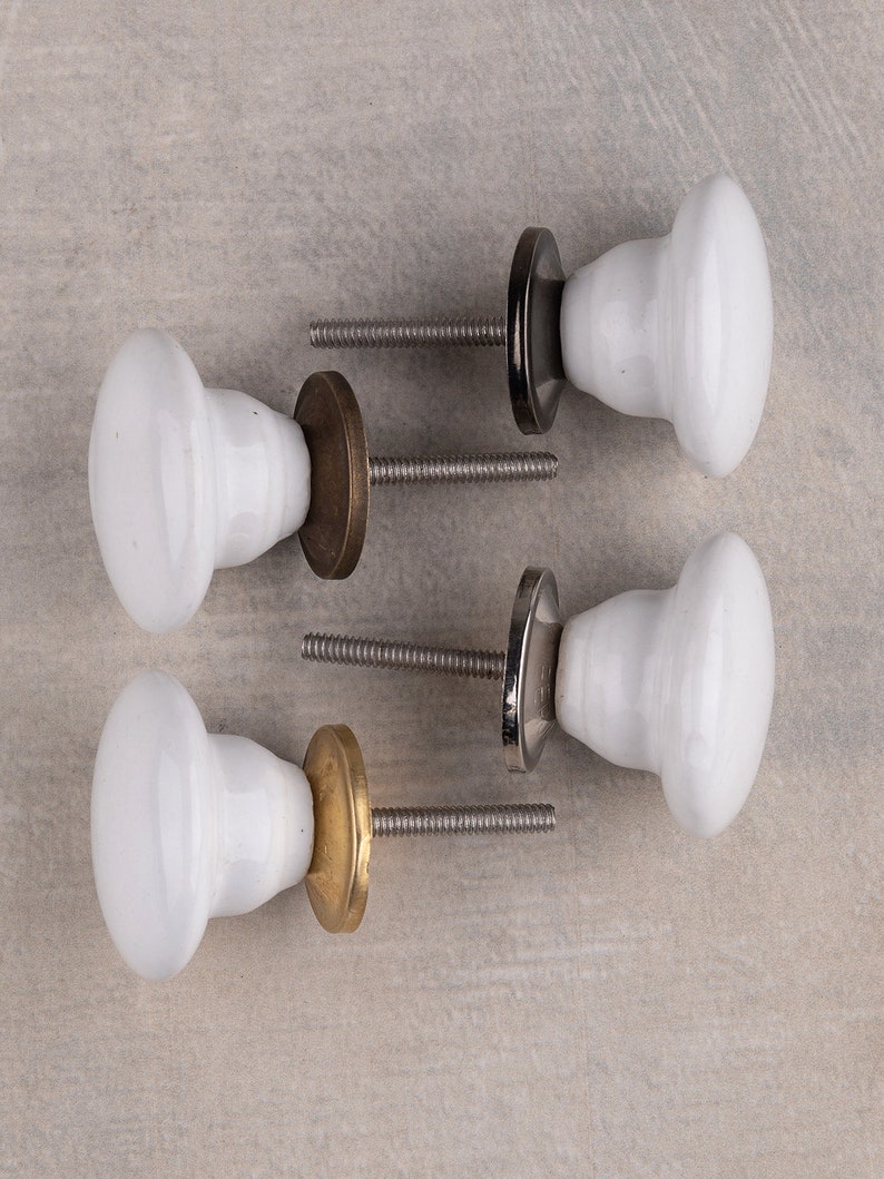 May include: Four white ceramic cabinet knobs with smooth, rounded tops and tapered bases. Each knob features a threaded metal bolt and a circular metal backplate in varying finishes, including dark bronze, polished silver, and brushed brass tones.